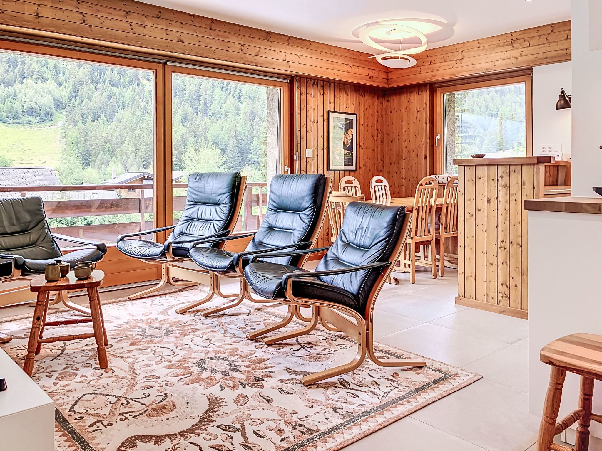 A room with a wooden interior, large windows, a dining area, and a seating arrangement with chairs and a table.