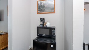 A compact kitchenette with a microwave and coffee maker, a framed landscape painting on the wall, and a small wooden cabinet.