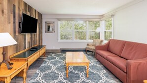 A living room with a wooden wall, a flat-screen TV, a sofa, a coffee table, a patterned rug, and a window with a view of trees. - Mountainside Inn 113 1 Bedroom Condo by Alpine Lodging Telluride (Telluride)