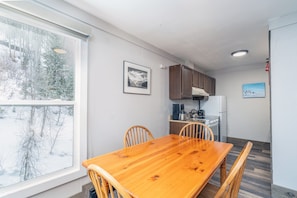 Uma sala de jantar com mesa e cadeiras de madeira, uma cozinha com armários e eletrodomésticos, e uma janela grande com vista para a neve.