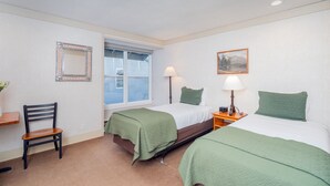 Quarto com duas camas, uma cadeira, uma mesa de cabeceira, um abajur, uma janela e um quadro na parede.