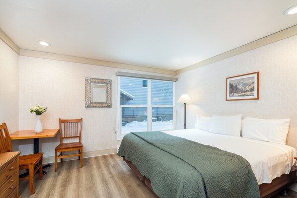 A bedroom with a bed, a desk with a chair, a vase with flowers, a framed mirror, and a window with a view of a snowy landscape. - Mountainside Inn 202 1 Bedroom Hotel Room by Alpine Lodging Telluride (Telluride)