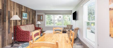 A dining area with a wooden table and chairs, a red patterned sofa, and a large window with a view of greenery.