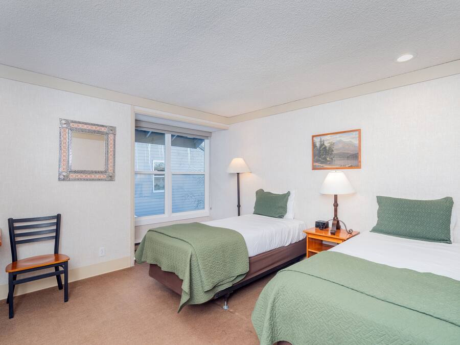 Une chambre avec deux lits, une chaise, une table de chevet, une lampe, une fenêtre et un tableau encadré au mur.