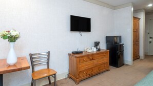 Une petite pièce bien éclairée, avec une commode en bois, un téléviseur fixé au mur, un réfrigérateur et une table à manger avec une chaise.