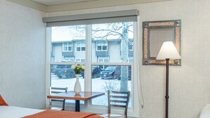 A hotel room with a large window, a bed with an orange bedspread, a small table with a vase of flowers, a chair, and a floor lamp. - Mountainside Inn 415 1 Bedroom Hotel Room by Alpine Lodging Telluride (Telluride)