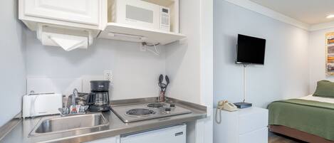 Una kitchenette compacta con microondas, tostadora, cafetera y un refrigerador pequeño. Un televisor de pantalla plana está montado en la pared sobre una cama con cubrecama verde.