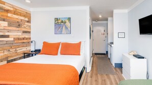 Hotellihuone, jossa on puinen sängynpääty, oranssilla tekstiilillä varustettu sänky, seinällä taulu, pieni keittiö ja seinälle asennettu televisio.