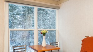 Une pièce avec une grande fenêtre, une table en bois, deux chaises et un lit avec un oreiller orange.