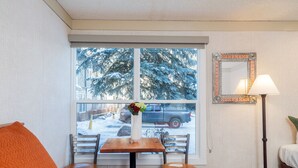 A hotel room with a bed, a table with a vase of flowers, a chair, a window with a view of snow-covered trees and a parked truck outside. - Mountainside Inn 304 1 Bedroom Hotel Room by Alpine Lodging Telluride (Telluride)