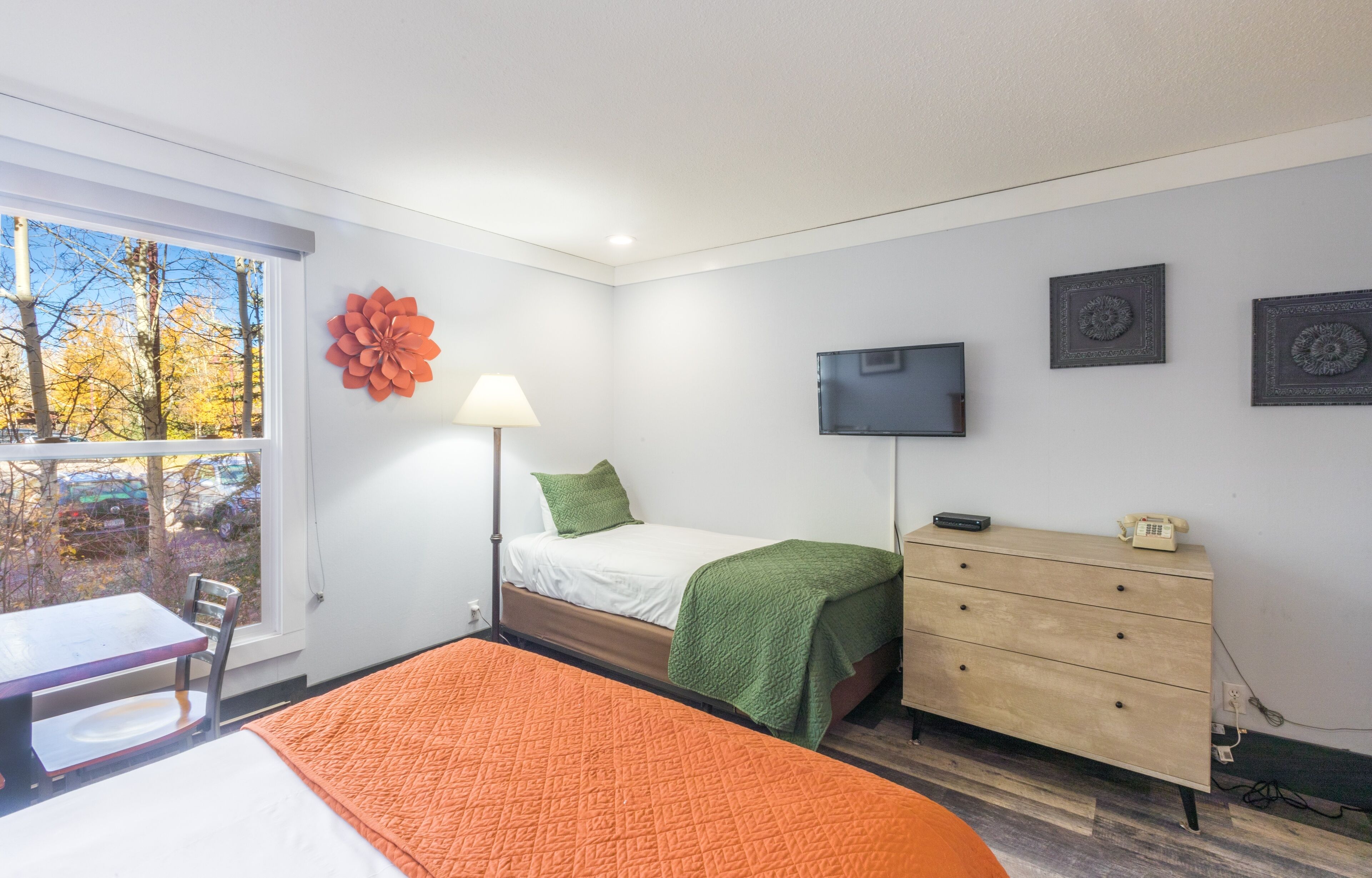 Un dormitorio con cama, escritorio, silla, cómoda, lámpara, televisor y ventana con vista a árboles y coches.