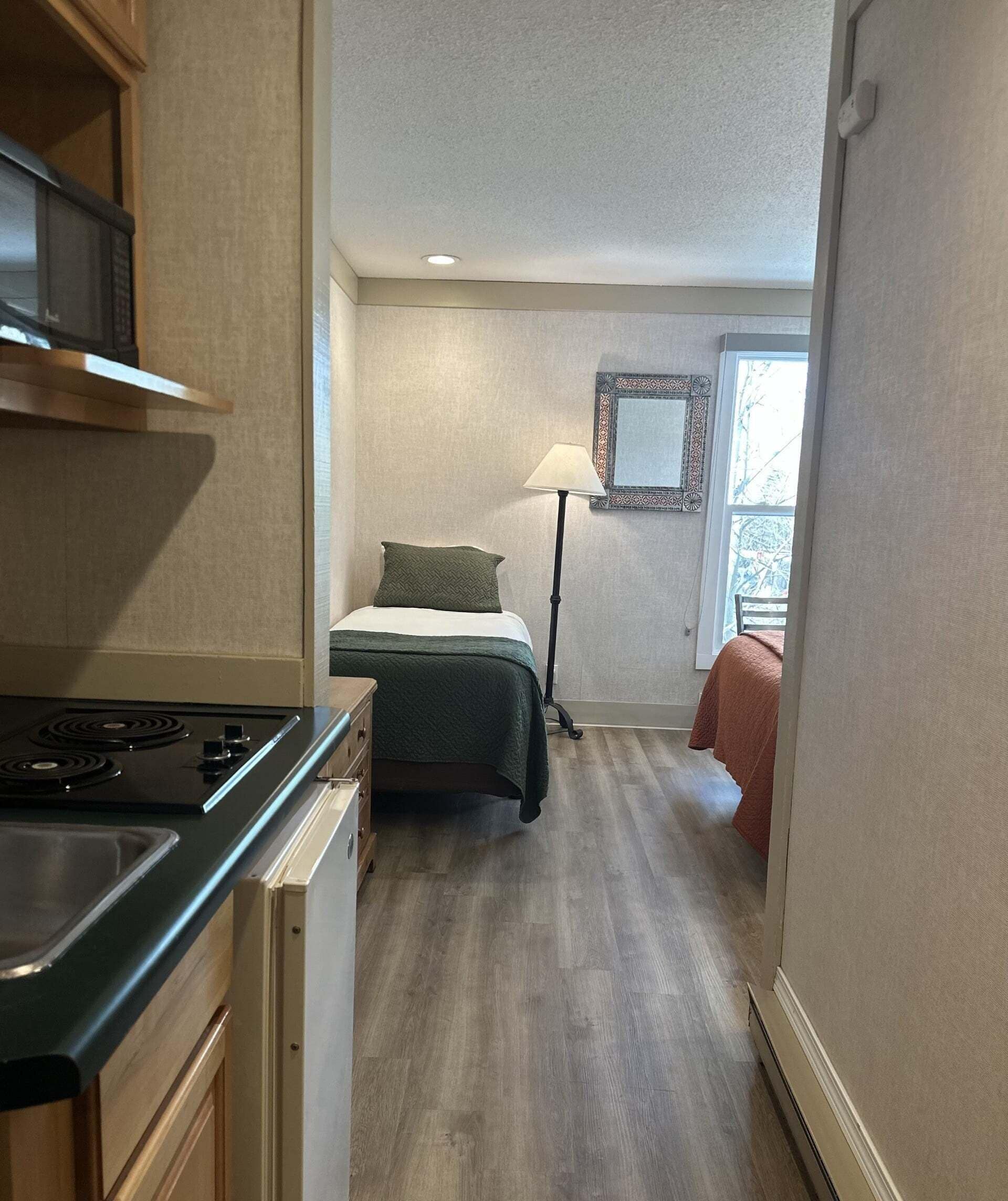 A compact kitchen with a stove and cabinets, a bed with a green blanket, a floor lamp, and a window with a view.