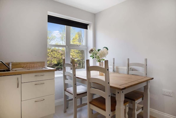 Una cocina con armarios blancos, una mesa de madera y sillas. Se ve una ventana con vistas a árboles.