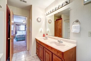 A bathroom with a double sink vanity, a large mirror with lights, a clock, and a towel rack.