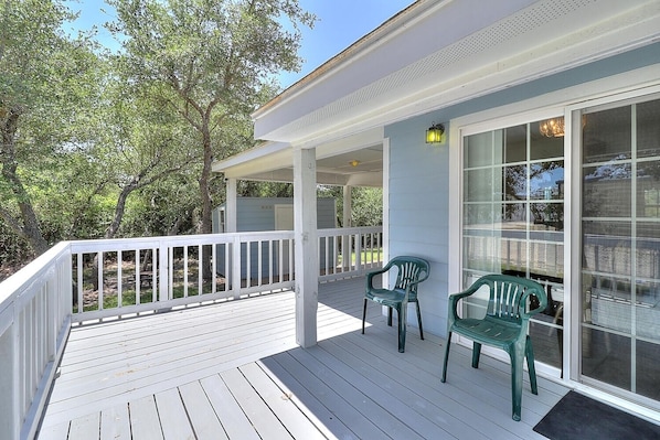 A covered porch with white railings, two green plastic chairs, and a large window with a view of trees and a building in the background.