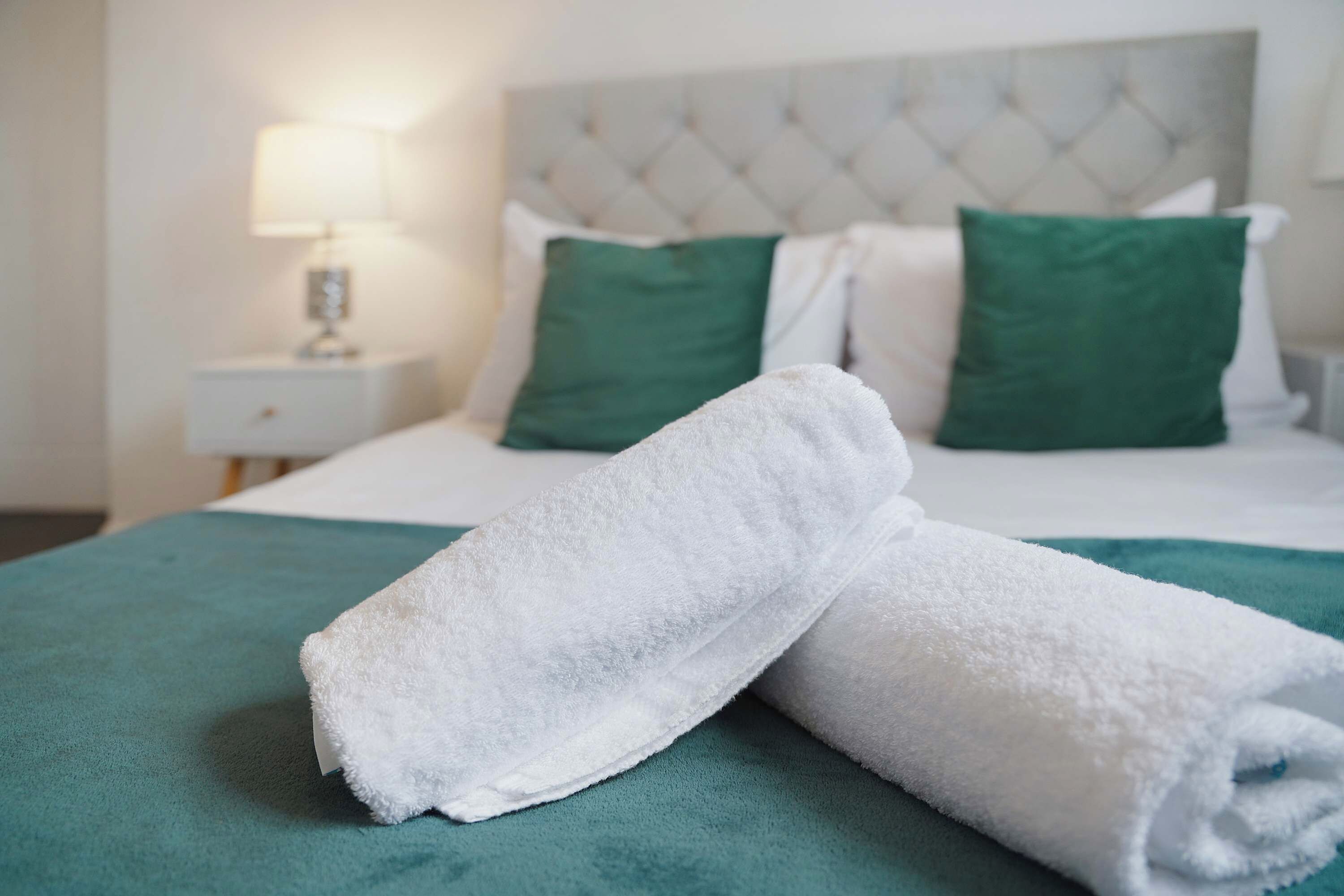A neatly made bed with white linens and green pillows, a white nightstand with a lamp, and a teal-colored bedspread.