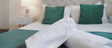 A neatly made bed with white linens and green pillows, a white nightstand with a lamp, and a teal-colored bedspread.