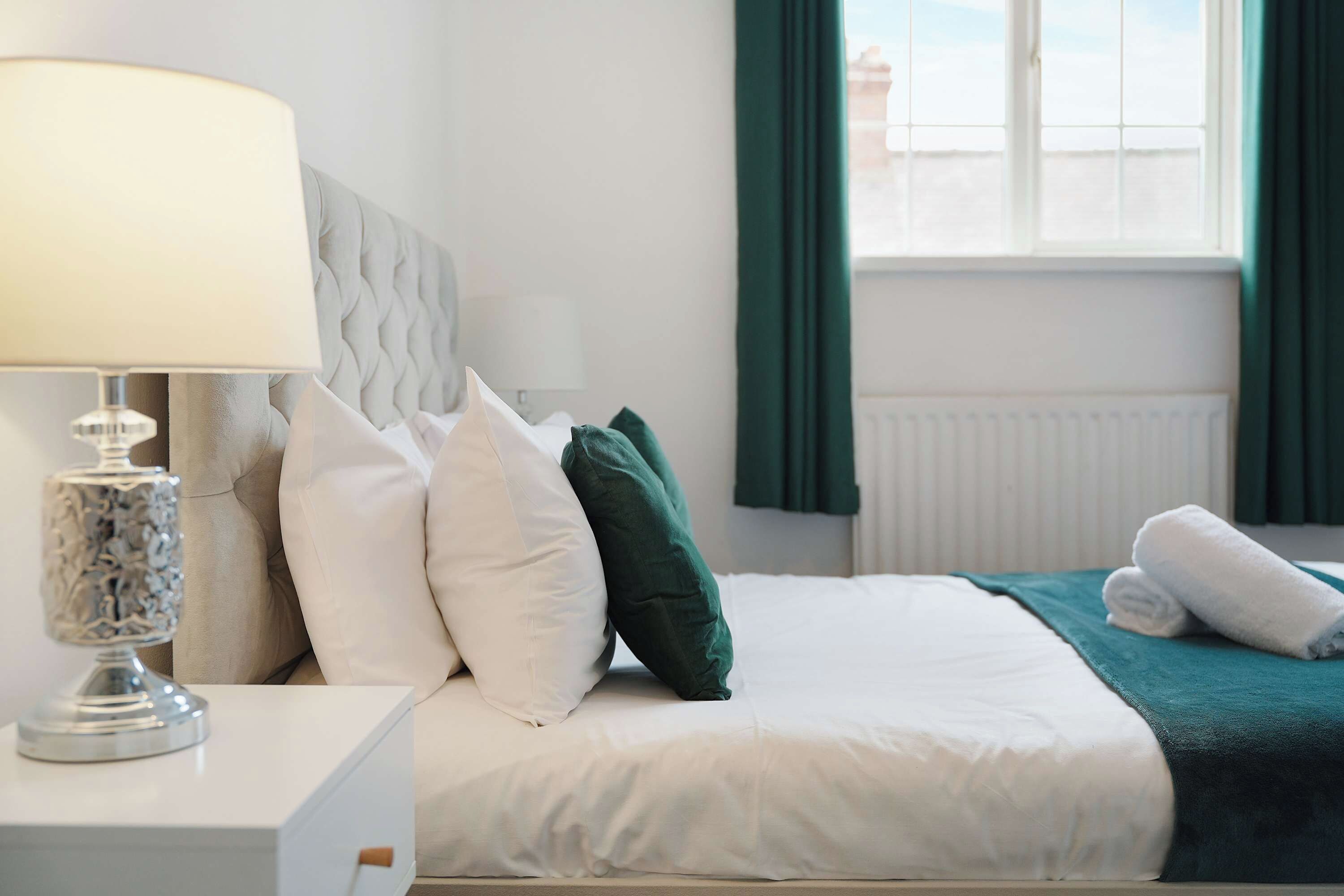 A neatly made bed with white bedding and a teal throw blanket, a bedside lamp with a textured base, and a window with green curtains.