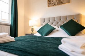 A neatly made bed with a green bedspread and pillows, a bedside lamp, and a window with curtains.