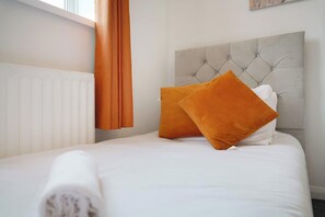 A neatly made bed with white bedding and two orange pillows, a grey tufted headboard, and a white radiator on the wall. - Cozy 3BR Stay in Houghton-le-Spring | Sleeps 5+WiFI (Hetton-le-Hole)