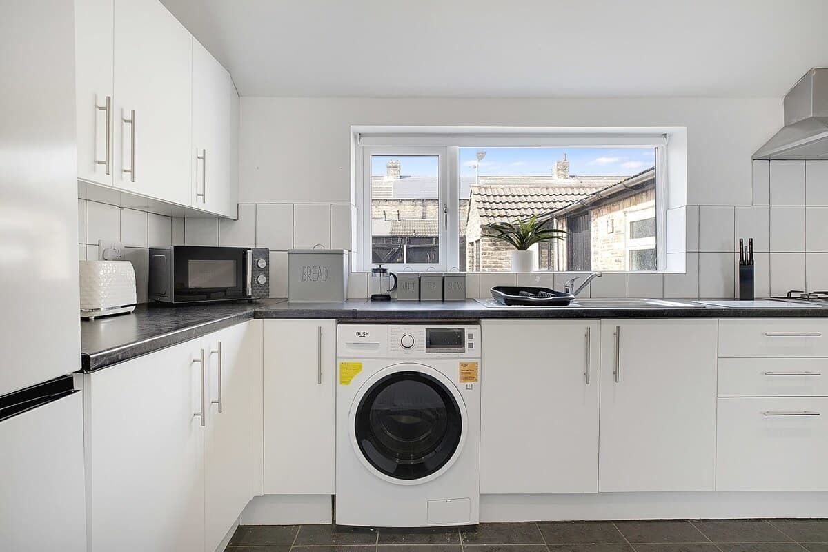 A modern kitchen with white cabinets, a microwave, a washing machine, and a window with a view of buildings.