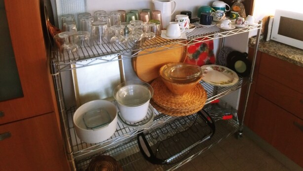 Une cuisine avec un étagère en métal où sont rangés différents objets en verre, un tableau accroché au mur et un four à micro-ondes posé sur le plan de travail.