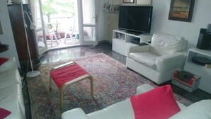 Un salon avec un canapé blanc, un pouf rouge, un téléviseur et une grande fenêtre avec des rideaux.