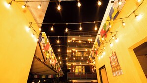 Un couloir bien éclairé, décoré de guirlandes lumineuses et de drapeaux colorés, qui mène à un bâtiment doté d’un balcon.