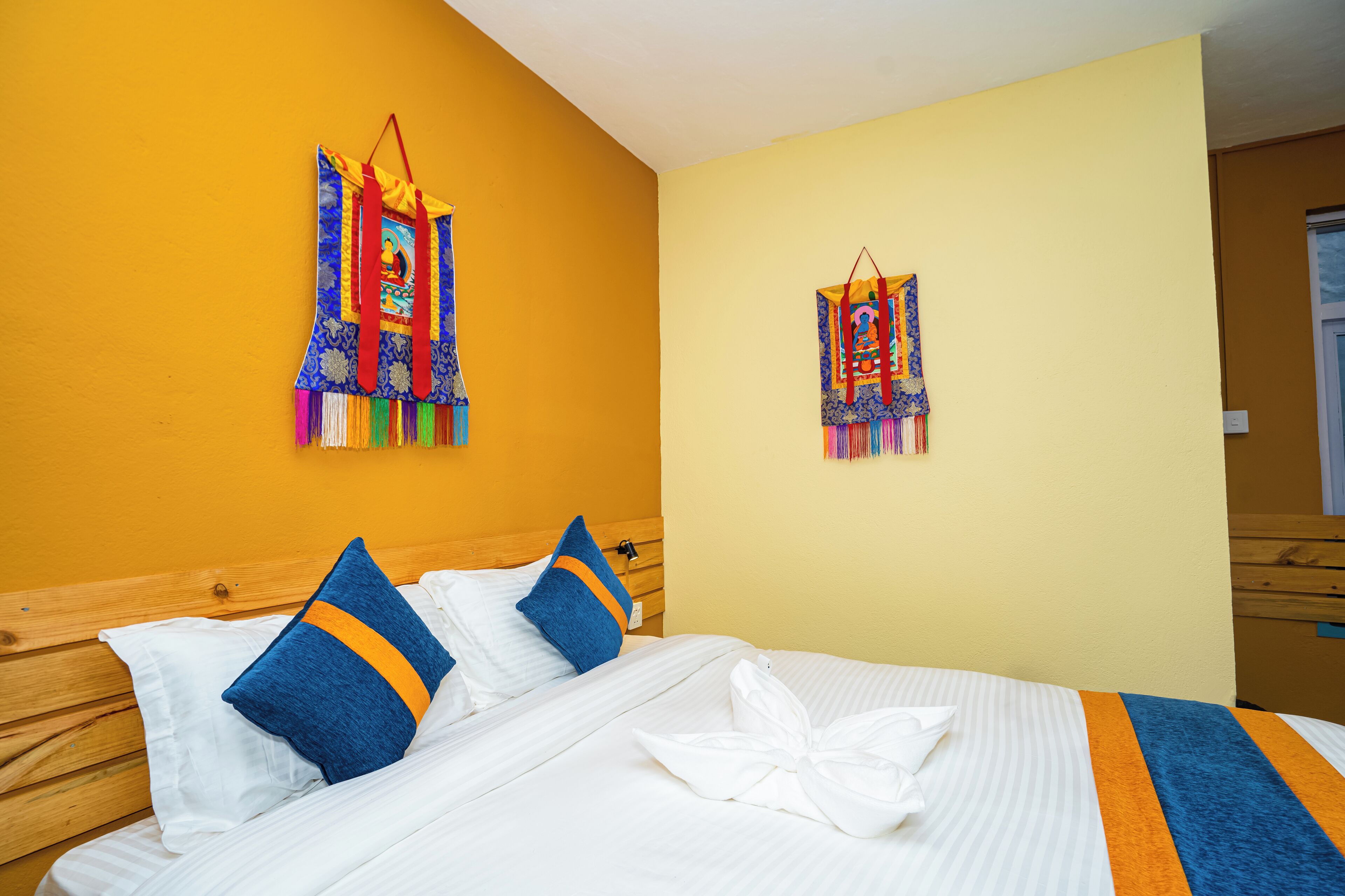 A hotel room with a wooden headboard, a bed with white bedding and blue pillows, and two colorful wall hangings.