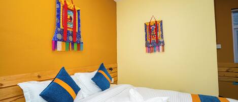 A hotel room with a wooden headboard, a bed with white bedding and blue pillows, and two colorful wall hangings.