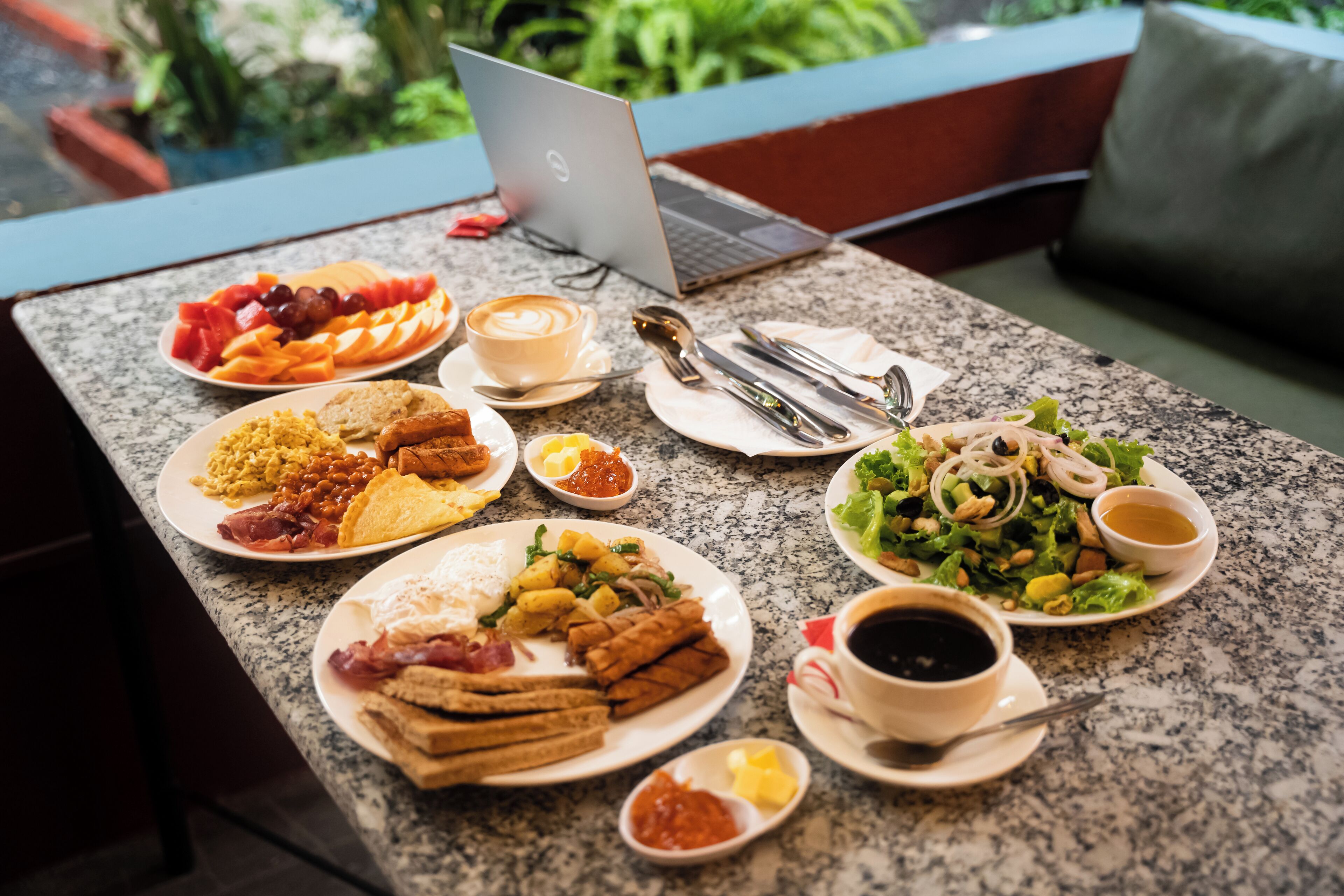 A breakfast buffet with various dishes, a laptop, and a coffee cup on a granite table.