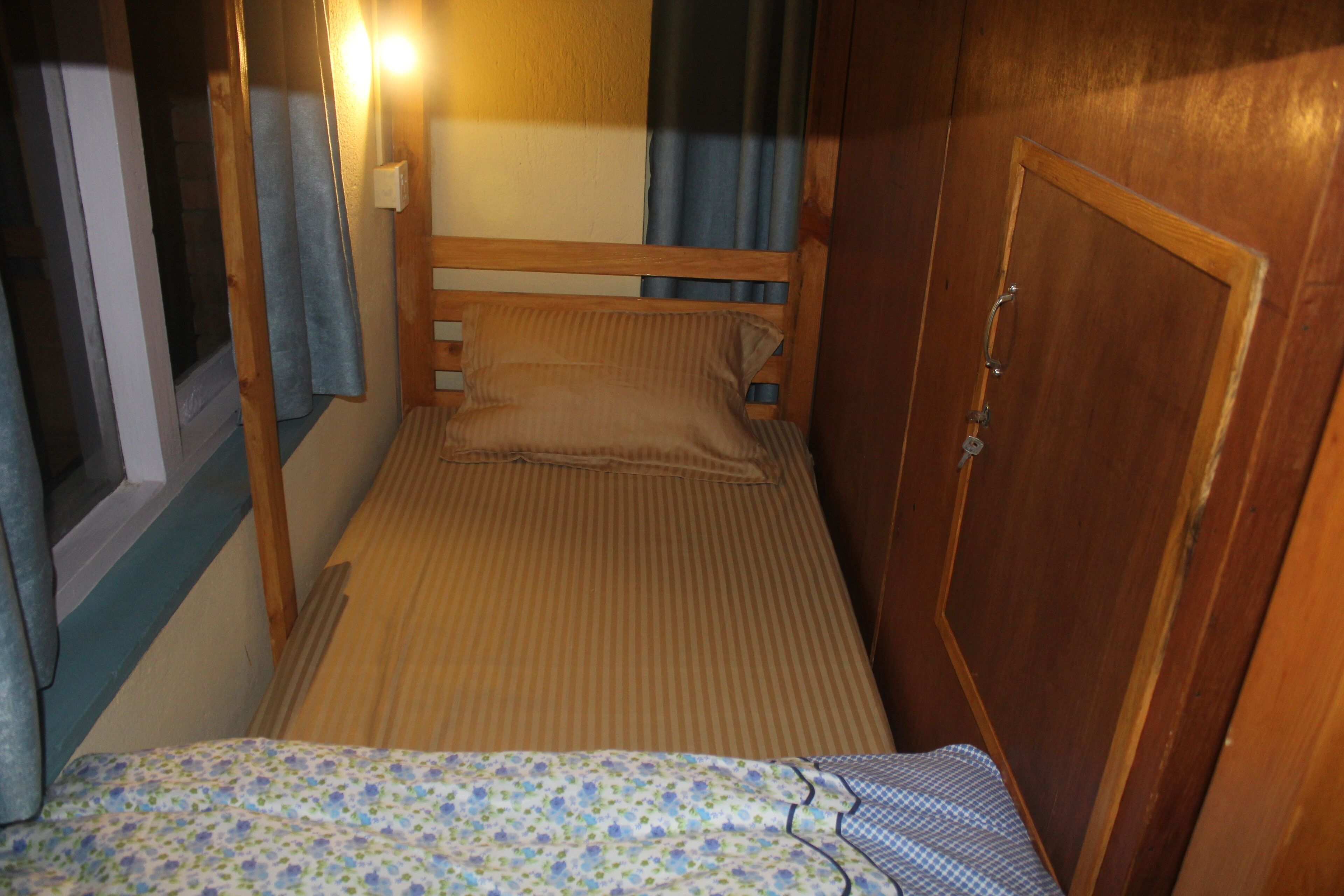 A small, narrow room with a single bed, a wooden wardrobe, and a window with curtains.