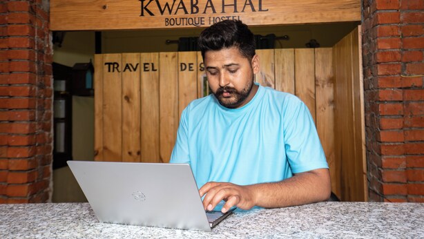 Une personne utilise un ordinateur portable à un comptoir, au-dessus duquel est affiché un panneau indiquant : « KWABAHAL BOUTIQUE HOSTEL ».