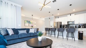 Un soggiorno moderno con un divano blu, un tavolino rotondo e un angolo cucina con pensili bianchi e sgabelli da bar.