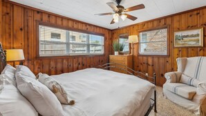 Un dormitorio con cama, una silla, ventilador de techo y dos ventanas.