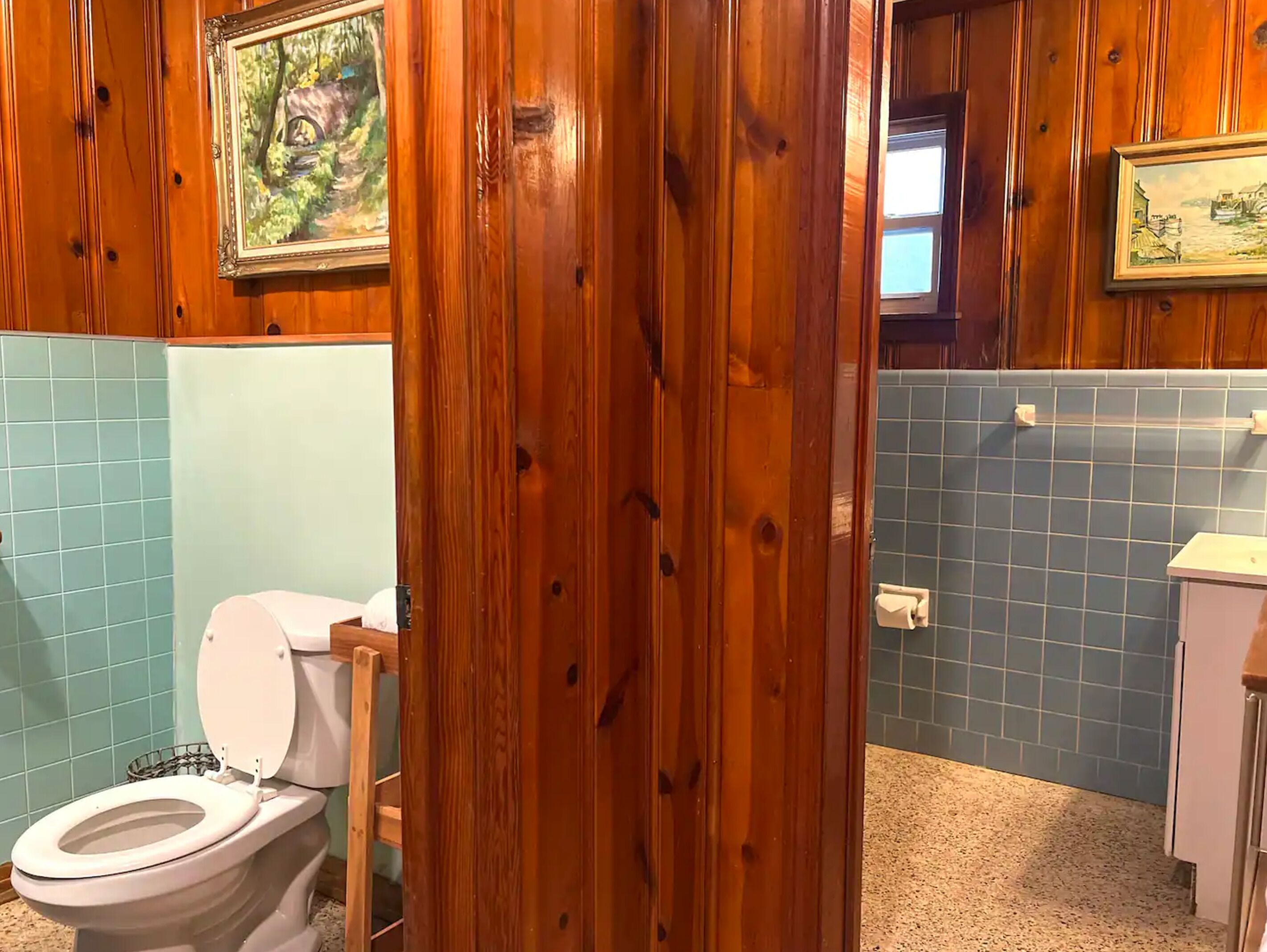 Une salle de bain aux murs lambrissés, équipée de toilettes, d’un lavabo et d’une fenêtre.