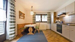 Een keuken met houten kastjes, een eettafel met stoelen, een blauw vloerkleed en een raam met gordijnen.