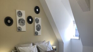 Une chambre avec un plafond en pente, un lit avec une literie blanche et des oreillers marron, une table de chevet avec une lampe et des œuvres d’art accrochées au mur.