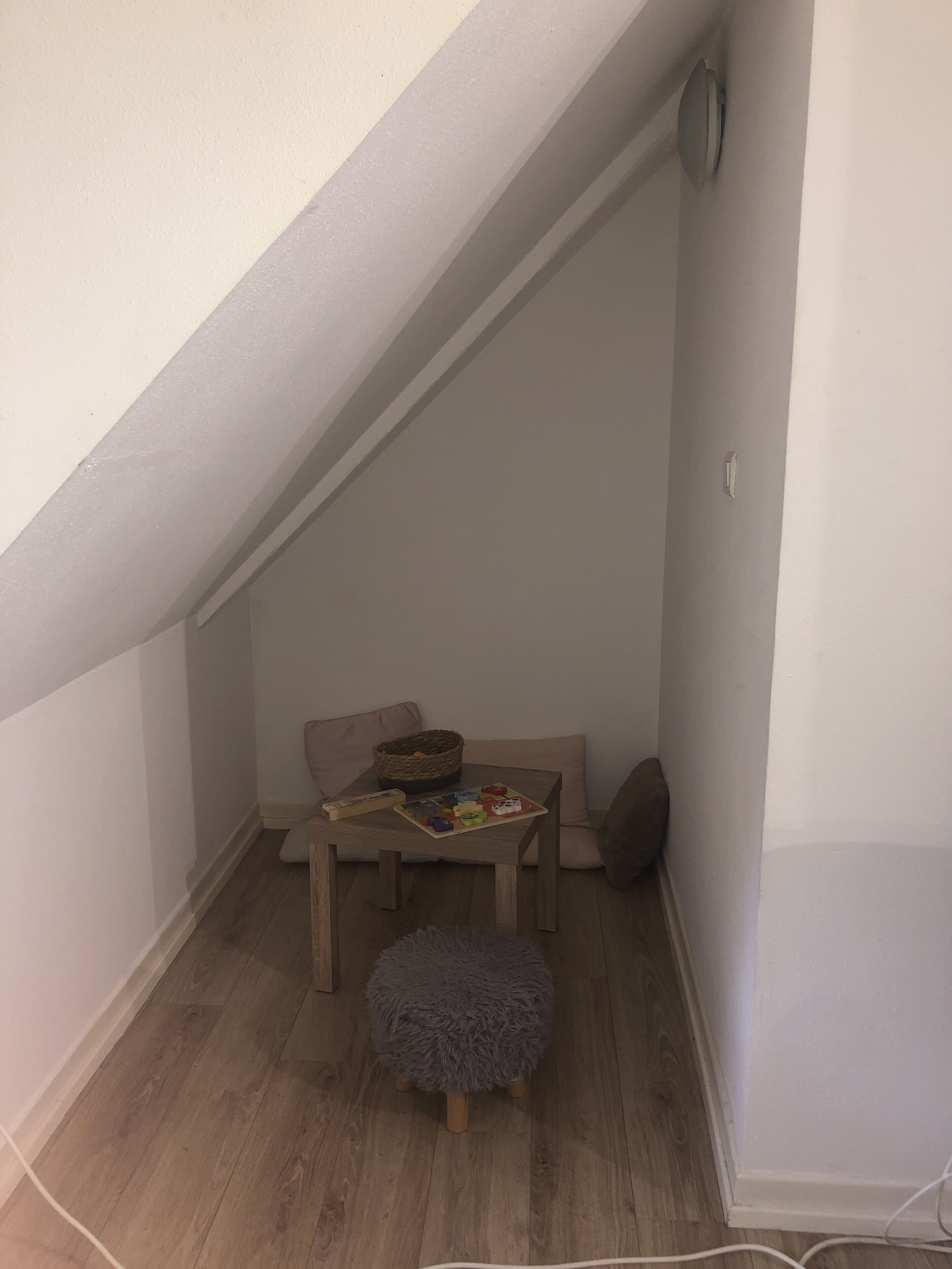 Ett litet, ombonat rum med ett sluttande tak, ett träbord med en korg och ett brädspel, en mjuk sittpuff och en brun stol.