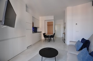 Een moderne woonkamer met een zwarte salontafel, een bank met blauwe kussens, een aan de muur bevestigde televisie, en een keuken met witte kasten en een houten deur.