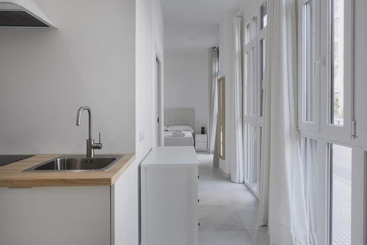 Um corredor estreito com uma área de cozinha à esquerda, uma cama com mesa de cabeceira no centro e uma janela à direita.