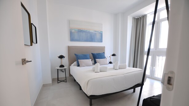 A modern hotel room with a large bed, a bedside table with a lamp, a painting on the wall, and a view of the outdoors through the window. - Capuchinos Cathedral (Málaga)