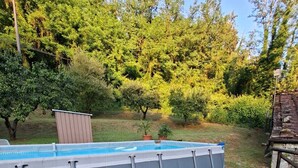 Una piscina elevada con estructura inflable gris, rodeada de césped artificial y árboles.