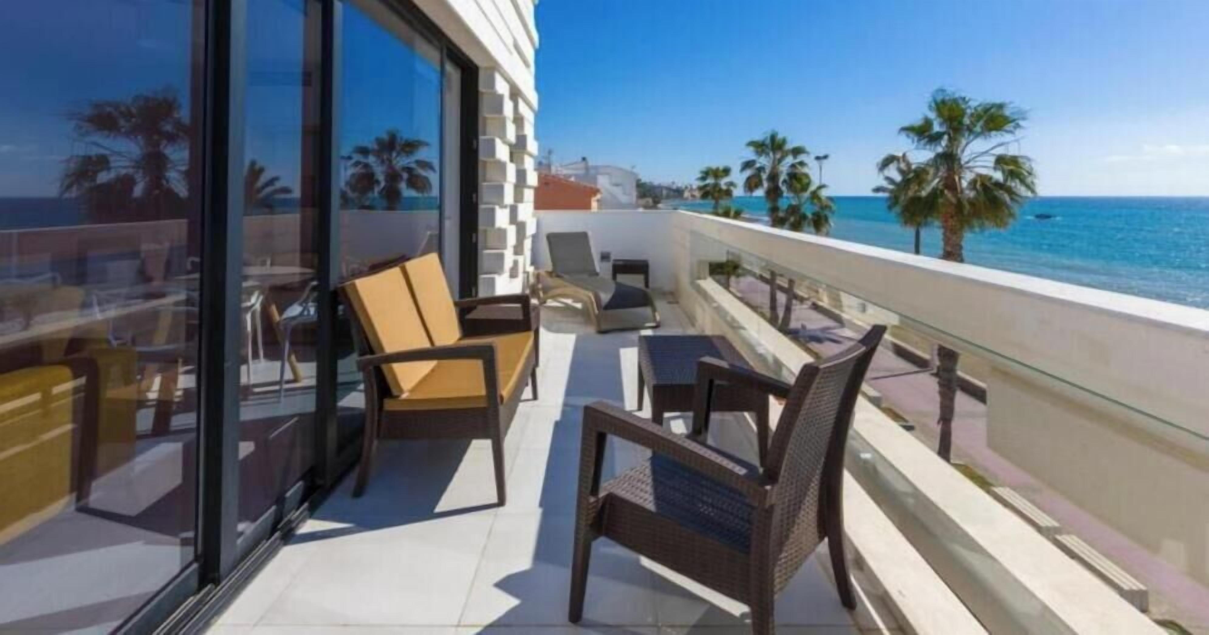 Een balkon met stoelen en een tafel, met uitzicht op een strand met palmbomen en de zee.