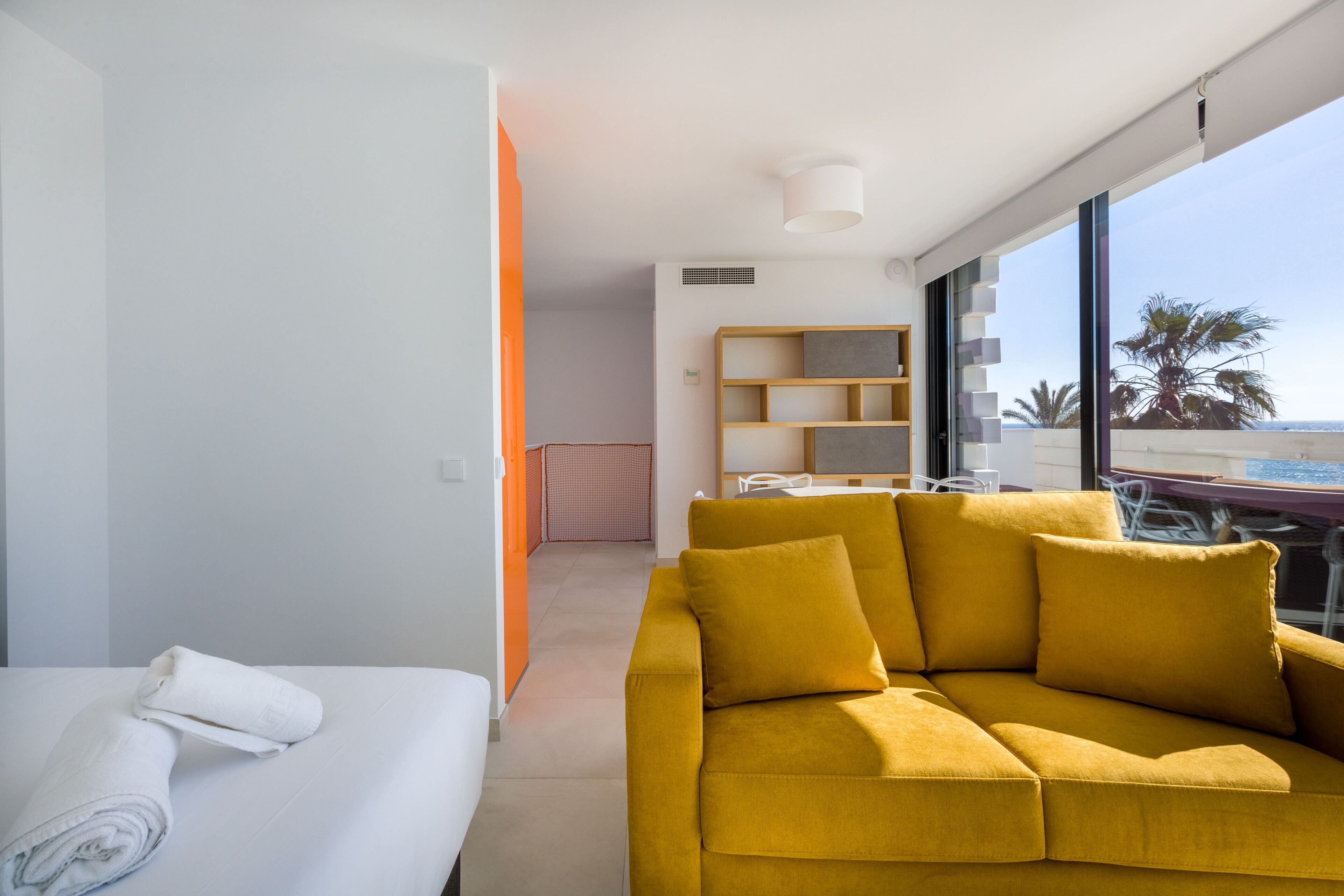 A modern hotel room with a yellow sofa, a white bed, and a large window offering a view of palm trees and the sea.