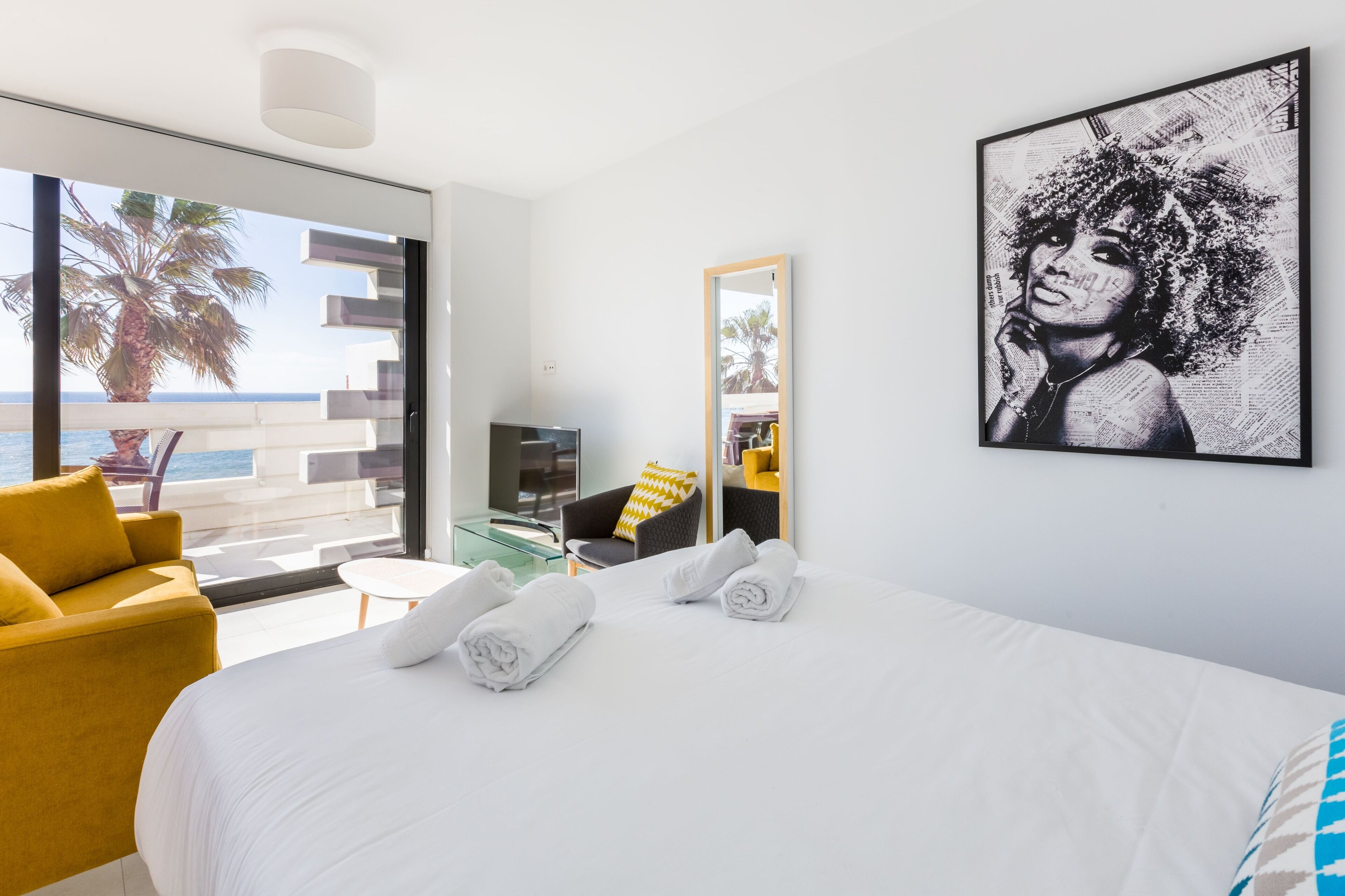 A hotel room with a large bed, a yellow armchair, a small table, and a large window with a view of the sea and palm trees.