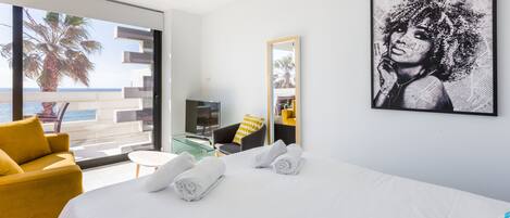 A hotel room with a large bed, a yellow armchair, a small table, and a large window with a view of the sea and palm trees.