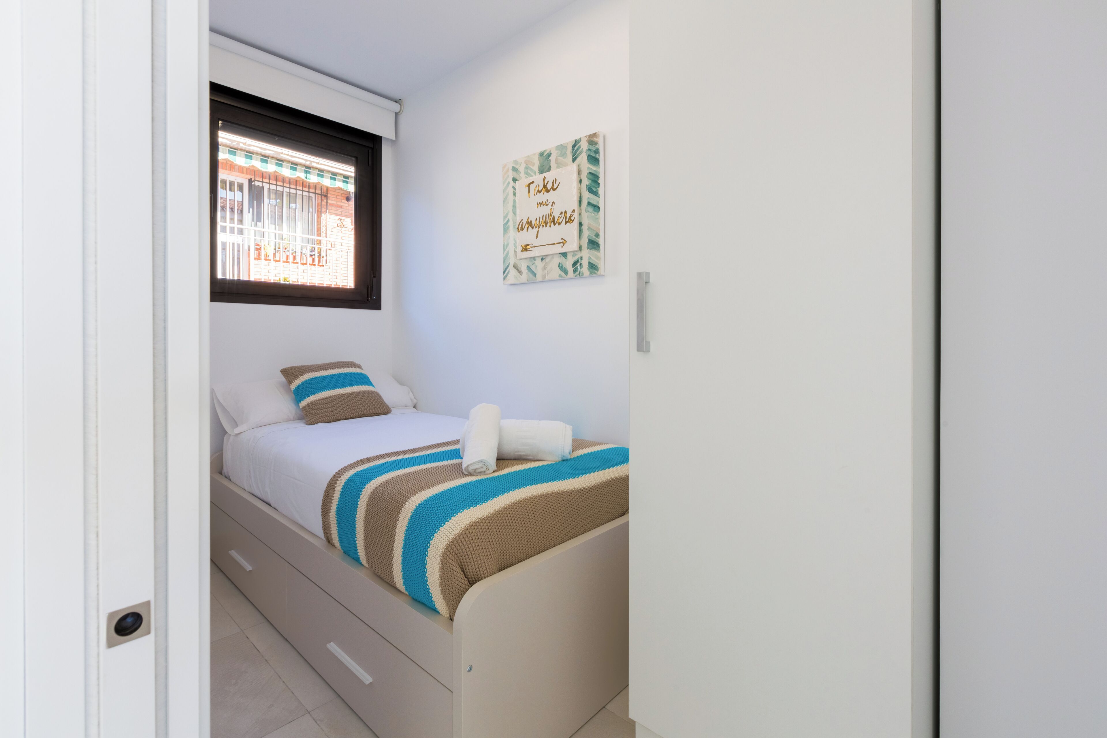 Une petite chambre à coucher, bien rangée, avec un lit, une fenêtre et une œuvre d’art murale inspirante.