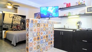 Una habitación pequeña y funcional con cama, encimera de cocina con azulejos, televisor y estante con artículos de aseo.