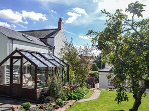 Una casa con un invernadero, un jardín con diversas plantas y un árbol de manzanas.